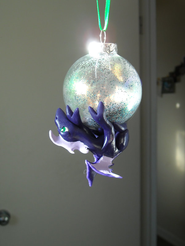 A purple polymer clay dragon hanging off he bottom of a round glass ornament full of glitter