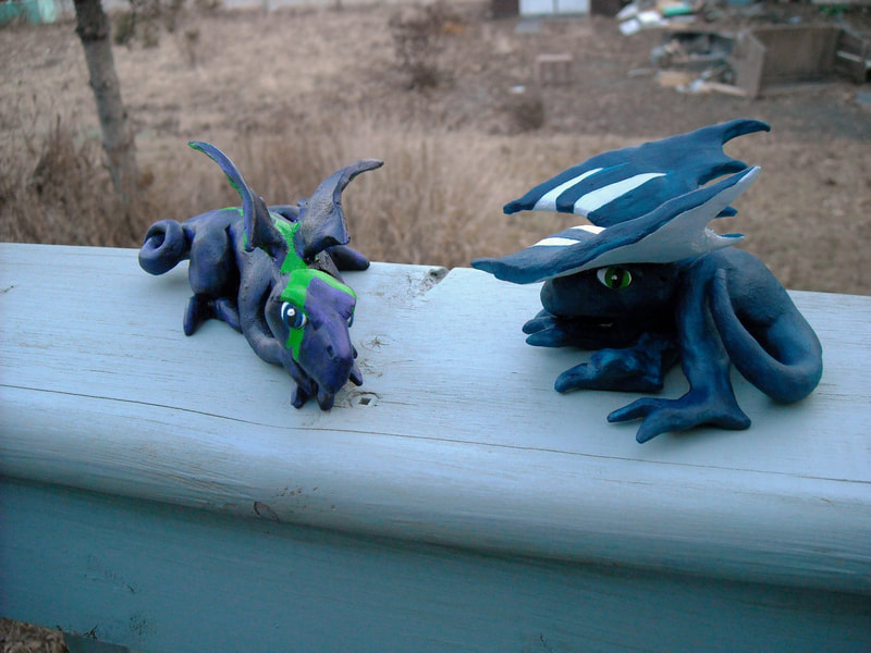 Two acrylic painted clay dragons, one is purple and green, the other is blue and silver. 